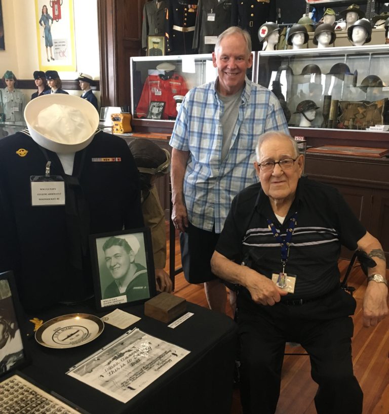 Two men pose together at a military museum exhibit, featuring a sailor's uniform and framed photographs, highlighting veterans' stories and memorabilia.